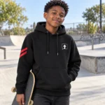 Teenage boy wearing a black SPEAK//UP pullover hoodie with an arrow chest patch and red sleeve stripes at a skatepark