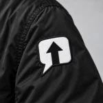 Close-up of the white speech-bubble arrow patch with black stitched edging on a black SPEAK//UP jacket sleeve