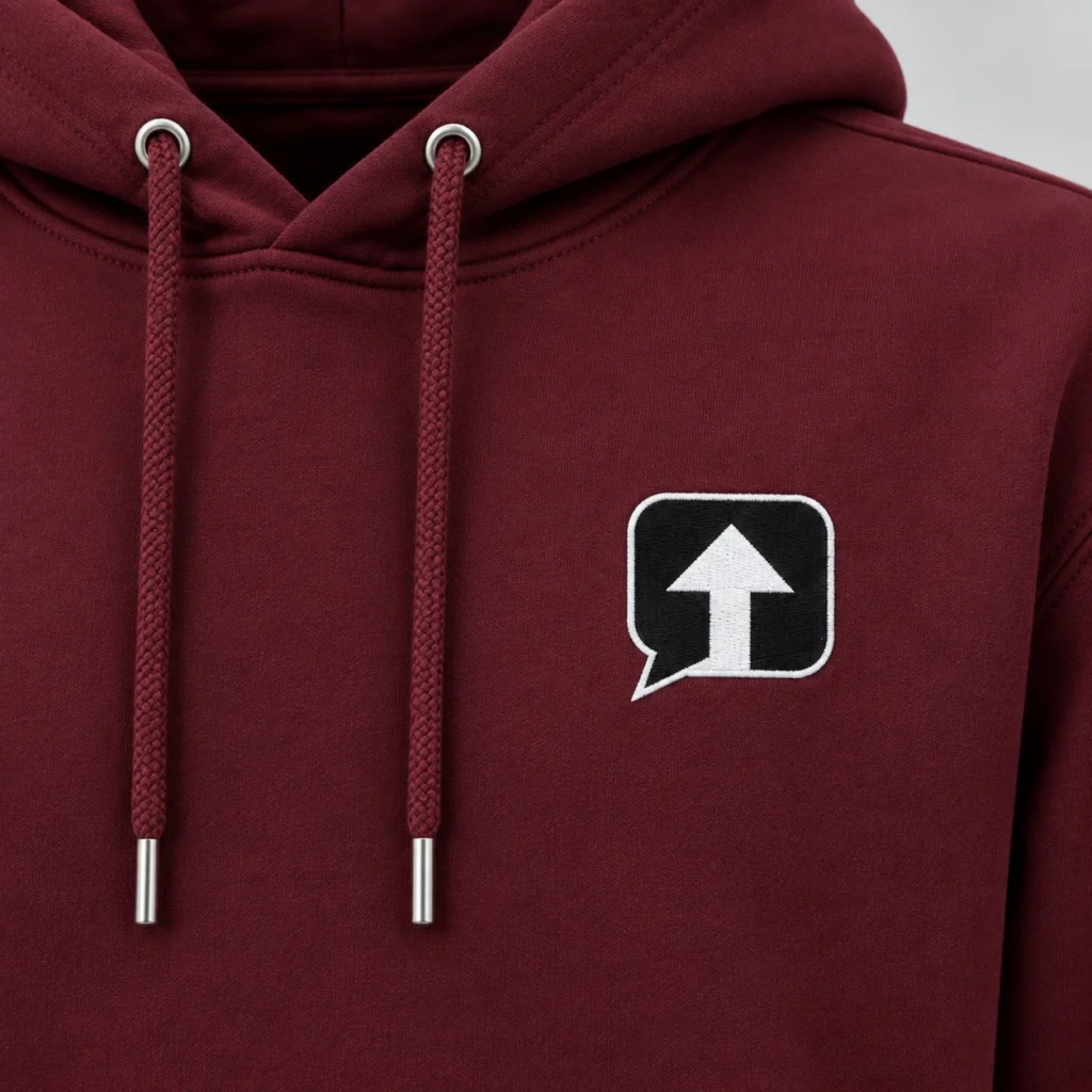 Close-up of the black speech-bubble patch with a white arrow and white stitched border on a burgundy SPEAK//UP hoodie