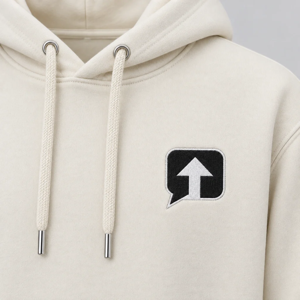 Close-up of the black speech-bubble patch with a white arrow and white stitched border on a milk ivory SPEAK//UP hoodie